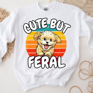 以下が含まれることがあります： レトロな夕焼けのデザインで、かわいい漫画の犬と「Cute But Feral」という文字が書かれた白いスウェットシャツです。
