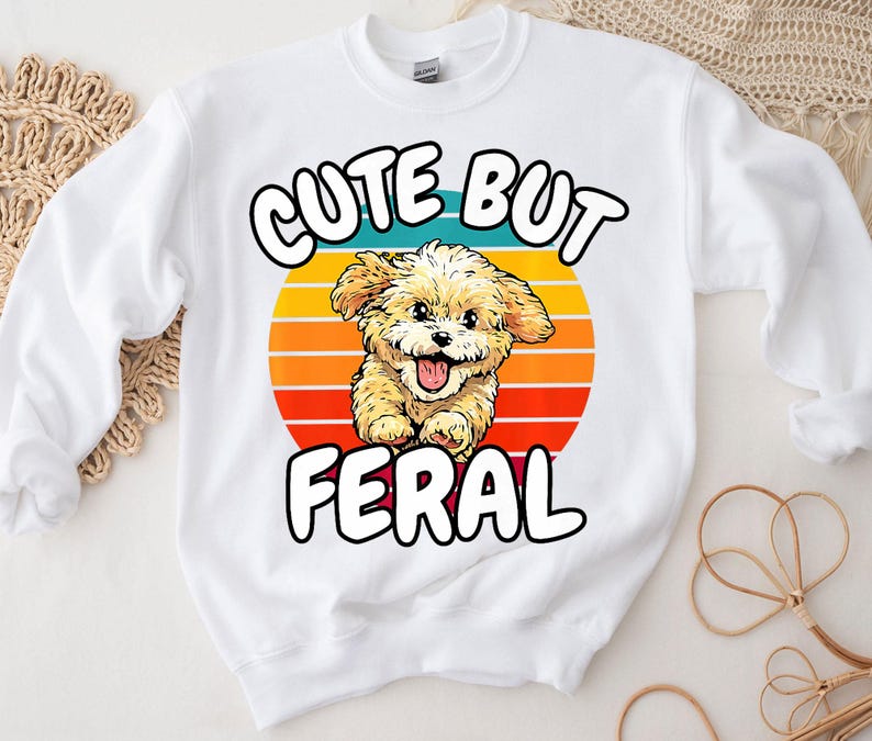 以下が含まれることがあります： レトロな夕焼けのデザインで、かわいい漫画の犬と「Cute But Feral」という文字が書かれた白いスウェットシャツです。
