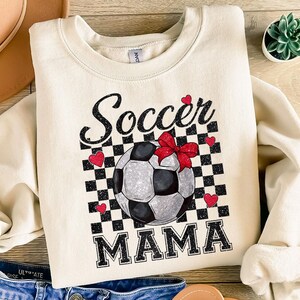 K&ouml;nnte beinhalten: Ein cremefarbener Pullover mit einem schwarz-wei&szlig;en Karomuster, das einen Fu&szlig;ball mit einer roten Schleife zeigt. Der Text "Soccer Mama" ist in schwarzen Buchstaben gedruckt.