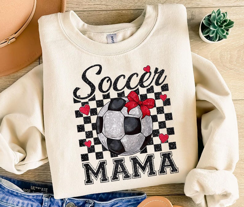 K&ouml;nnte beinhalten: Ein cremefarbener Pullover mit einem schwarz-wei&szlig;en Karomuster, das einen Fu&szlig;ball mit einer roten Schleife zeigt. Der Text "Soccer Mama" ist in schwarzen Buchstaben gedruckt.
