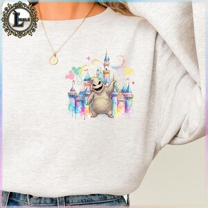 Puede incluir: Una sudadera gris claro con un gr&aacute;fico colorido de un personaje de dibujos animados frente a un castillo. El castillo est&aacute; representado con tonos azules, rosas y amarillos, con un arco&iacute;ris y estrellas. El personaje es de color beige claro con una cara cosida.