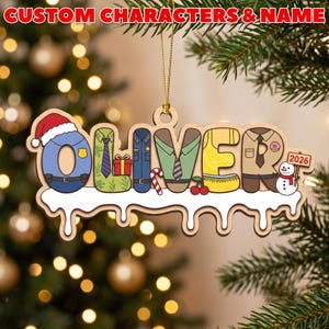 May include: A wooden Christmas ornament featuring the name OLIVER spelled out in colorful, character-themed letters. The design includes a Santa hat, a gift, a candy cane, cherries, a donut, and a snowman holding a sign that reads 2026.