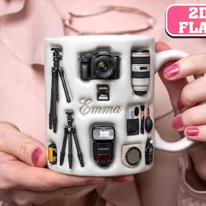 May include: A white ceramic mug featuring a 2D flat graphic print of photography equipment including a camera, two tripods, a lens, a flash, a memory card, a film roll, and various cleaning tools. The name Emma is printed in script in the center.