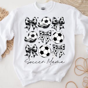 K&ouml;nnte beinhalten: Wei&szlig;er Pullover mit einem schwarz-wei&szlig;en Design aus Fu&szlig;b&auml;llen, Schleifen und dem Text "Soccer Mama".