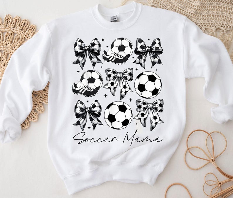 K&ouml;nnte beinhalten: Wei&szlig;er Pullover mit einem schwarz-wei&szlig;en Design aus Fu&szlig;b&auml;llen, Schleifen und dem Text "Soccer Mama".