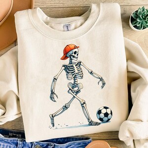 K&ouml;nnte beinhalten: Ein cremefarbener Sweatshirt mit einem Aufdruck eines Skeletts, das eine rote Baseballkappe tr&auml;gt und einen Fu&szlig;ball tritt. Das Skelett tr&auml;gt ein wei&szlig;es Hemd und schwarze Shorts.