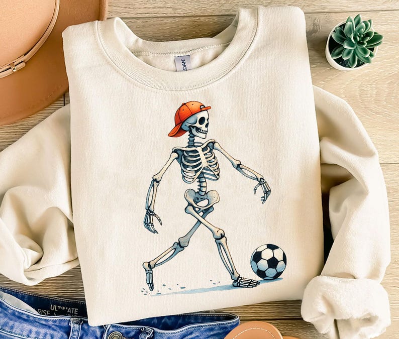 K&ouml;nnte beinhalten: Ein cremefarbener Sweatshirt mit einem Aufdruck eines Skeletts, das eine rote Baseballkappe tr&auml;gt und einen Fu&szlig;ball tritt. Das Skelett tr&auml;gt ein wei&szlig;es Hemd und schwarze Shorts.