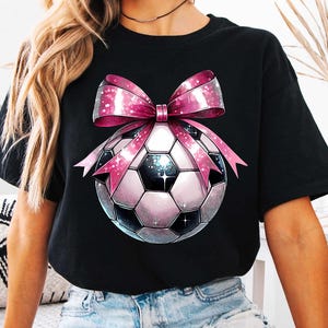 K&ouml;nnte beinhalten: Schwarzes T-Shirt mit einer rosa Schleife und einem Fu&szlig;ball-Motiv. Der Fu&szlig;ball ist wei&szlig; mit schwarzen Feldern und einem glitzernden Design.