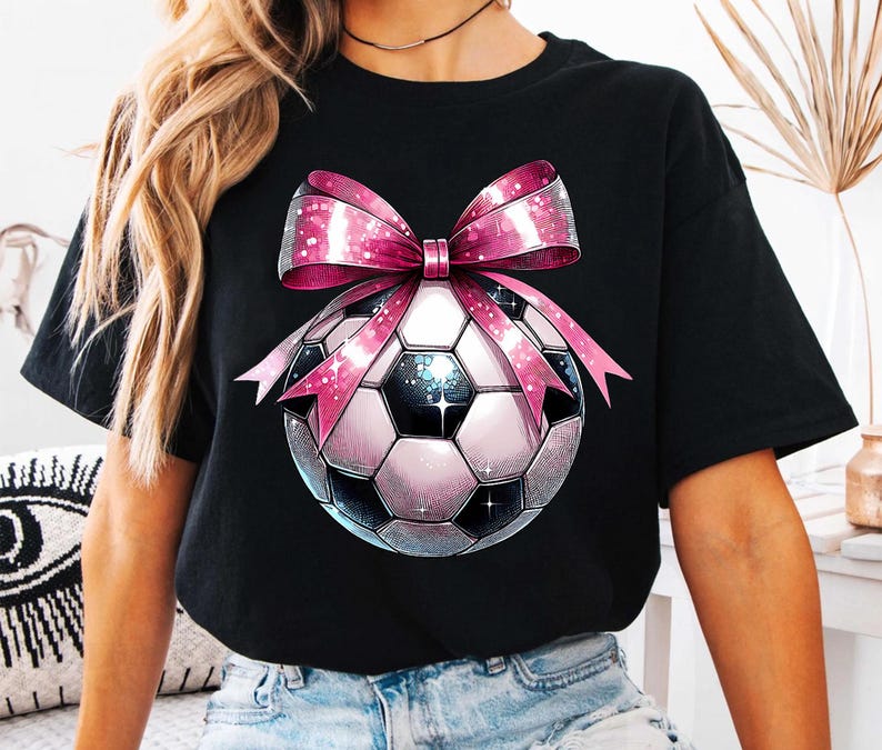 K&ouml;nnte beinhalten: Schwarzes T-Shirt mit einer rosa Schleife und einem Fu&szlig;ball-Motiv. Der Fu&szlig;ball ist wei&szlig; mit schwarzen Feldern und einem glitzernden Design.