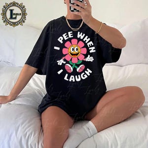 May include: Black t-shirt featuring a smiling flower graphic with the text "I PEE WHEN I LAUGH" in white. The flower has pink petals, a yellow center, and green stem and leaves. The shirt is a casual, oversized style.