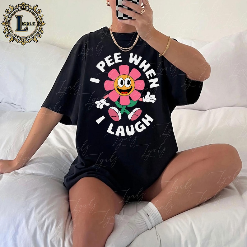 May include: Black t-shirt featuring a smiling flower graphic with the text "I PEE WHEN I LAUGH" in white. The flower has pink petals, a yellow center, and green stem and leaves. The shirt is a casual, oversized style.