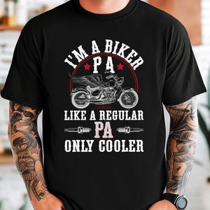 Puede incluir: Camiseta negra con un dise&ntilde;o gr&aacute;fico blanco que dice "I'm a biker PA like a regular PA only cooler". El gr&aacute;fico tambi&eacute;n incluye una motocicleta y buj&iacute;as.