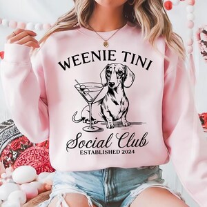 以下が含まれることがあります： 「Weenie Tini Social Club Established 2024」という文字と、マルティーニグラスから飲んでいるダックスフントの白黒イラストが描かれたピンク色のスウェットシャツです。