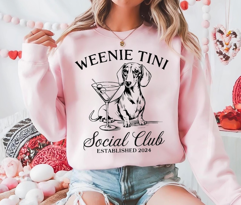 以下が含まれることがあります： 「Weenie Tini Social Club Established 2024」という文字と、マルティーニグラスから飲んでいるダックスフントの白黒イラストが描かれたピンク色のスウェットシャツです。