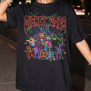 May include: A black short-sleeve t-shirt featuring a colorful graphic print with the text DISNEY 2026 at the top. The design includes characters from The Nightmare Before Christmas, a haunted castle, bats, and a tombstone with TRICK OR TREAT text.