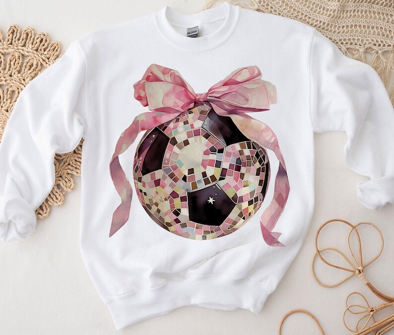 K&ouml;nnte beinhalten: Wei&szlig;er Pullover mit einer rosa Schleife und einem Discokugel-Fu&szlig;ball-Motiv.