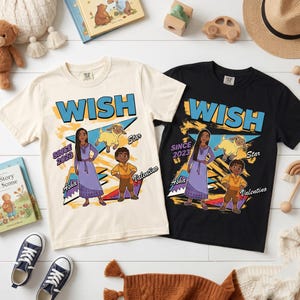 May include: Two short-sleeve t-shirts, one cream and one black, featuring a graphic print with the text WISH, SINCE 2023, Asha, Star, and Valentino. The design includes illustrations of the characters Asha, a goat named Star, and Valentino.