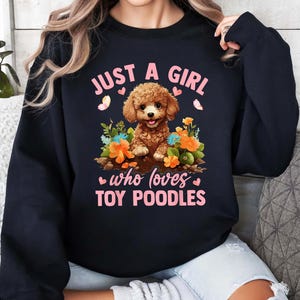 以下が含まれることがあります： ピンクの文字で「Just a girl who loves toy poodles」と書かれた黒いスウェットシャツ。テキストの中央には、ピンクの花を持った茶色のプードルの漫画が描かれています。