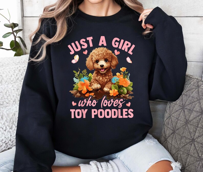 以下が含まれることがあります： ピンクの文字で「Just a girl who loves toy poodles」と書かれた黒いスウェットシャツ。テキストの中央には、ピンクの花を持った茶色のプードルの漫画が描かれています。