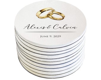 May include: A stack of round white coasters with a gold ring design and the text "Alexis & Calvin June 9, 2029" printed on the top coaster.