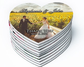 May include: A stack of heart-shaped wedding favors with a photo of a bride and groom in a sunflower field. The text on the favors reads "Stephanie & John" and "Thank You May 8, 2021".