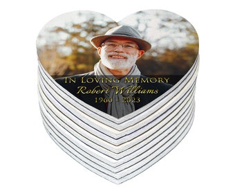 May include: Heart-shaped memorial keepsake with a photo of a smiling man wearing a hat and glasses. The text "IN LOVING MEMORY Robert Williams 1960 - 2023" is printed in gold. The edges have a striped pattern.