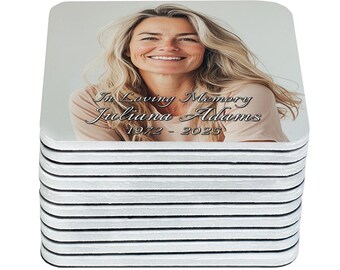 May include: A stack of square coasters featuring a photograph of a smiling woman with blonde hair. The coasters have the text "In Loving Memory Juliana Adams 1972 - 2025" printed on them. The coasters are white with black edges.