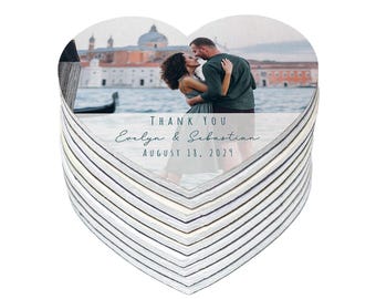 May include: Heart-shaped wedding favor with a photo of a couple kissing in front of a building. The text "THANK YOU Evelyn & Sebastian AUGUST 18, 2029" is printed on the front. The favors are stacked.
