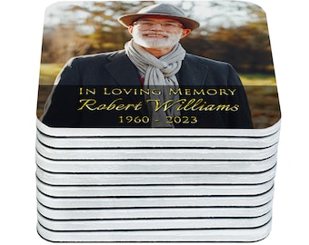 May include: A stack of 12 square coasters with a photo of a man wearing a hat and scarf. The coasters have a black background with gold text that reads "In Loving Memory Robert Williams 1960 - 2023".