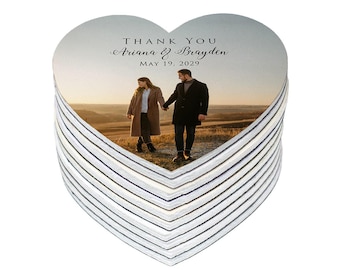 May include: A stack of heart-shaped coasters. Each coaster features a photo of a couple holding hands in a field, with the text "THANK YOU Ariana & Brayden MAY 19, 2029" printed at the top. The coasters have a white and gray striped edge.