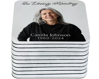 May include: A stack of square coasters featuring a photograph of a woman with gray hair, wearing a black top. The coasters have the text "In Loving Memory" and the name "Camila Johnson 1960-2024". The coasters have a white background.