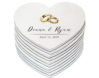 May include: A stack of white heart-shaped wedding favors with gold rings and the text "Diana & Ryan" and "MAY 12, 2029".