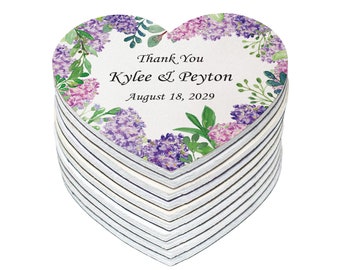 May include: A stack of heart-shaped wedding favors with a floral design. The favors are white with purple and pink flowers and green leaves. The text on the favors reads "Thank You Kylee & Peyton August 18, 2029".