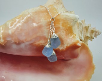 May include: A silver chain necklace with three light blue sea glass pendants wrapped in silver wire. The necklace is displayed on a pink seashell.