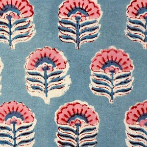 May include: A close-up of a fabric featuring a repeating floral design. The flowers have pink petals, blue centres, and white and blue stems, set against a light blue background. The design is reminiscent of block printing.