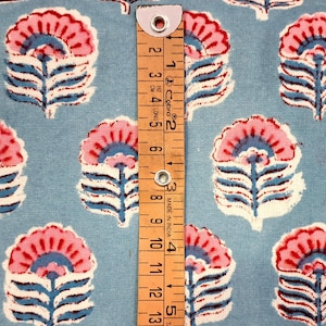 May include: Pale blue fabric featuring a repeating floral design in pink, white, and blue. A wooden ruler with black markings and the text "MADE IN INDIA" is placed on the fabric. The ruler measures up to 15 cm.