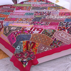 Op de afbeelding: Een kleurrijk patchwork-deken met een roze rand. Het deken is gemaakt van verschillende stoffen met verschillende patronen en kleuren, waaronder rood, blauw, groen en geel. Het deken ligt op een bed met een wit kussen.
