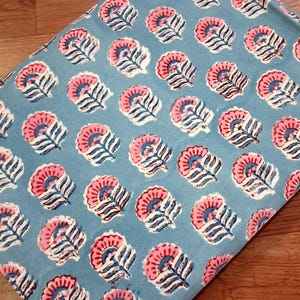 May include: A folded piece of fabric with a repeating floral pattern. The flowers are pink with blue centers and white accents, set against a light blue background. The fabric is likely cotton or linen.