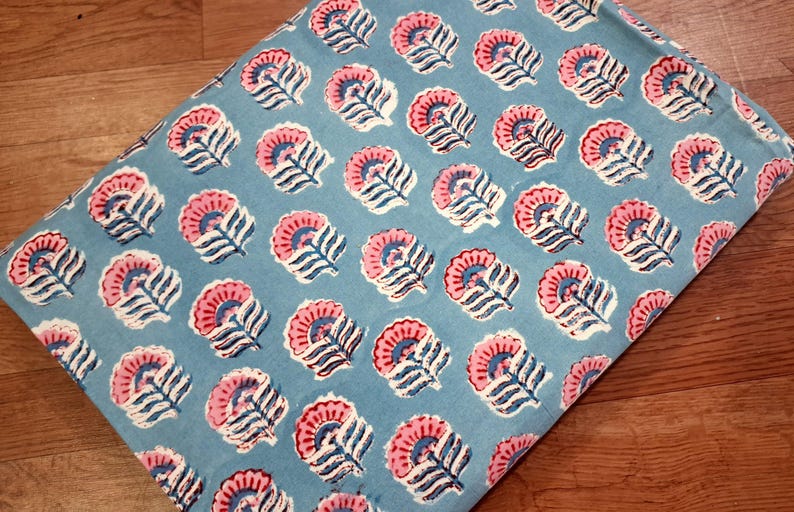 May include: A folded piece of fabric with a repeating floral pattern. The flowers are pink with blue centers and white accents, set against a light blue background. The fabric is likely cotton or linen.
