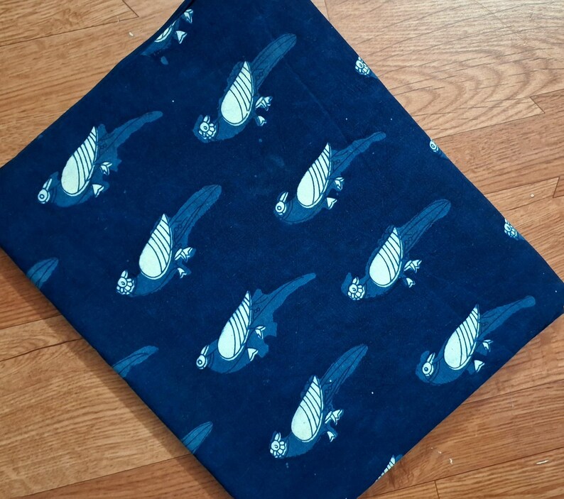 May include: A folded piece of dark blue fabric featuring a repeating pattern of stylised parrots. The birds are rendered in a lighter shade of blue with white wing details, arranged in a diagonal orientation across the entire surface of the textile.