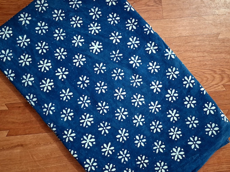 Pode incluir: Uma pe&ccedil;a de tecido retangular com um fundo azul profundo e um padr&atilde;o repetitivo de pequenas flores brancas de seis p&eacute;talas dispostas em grade. O tecido tem uma apar&ecirc;ncia plana e consistente com um contraste claro entre o azul e o branco.