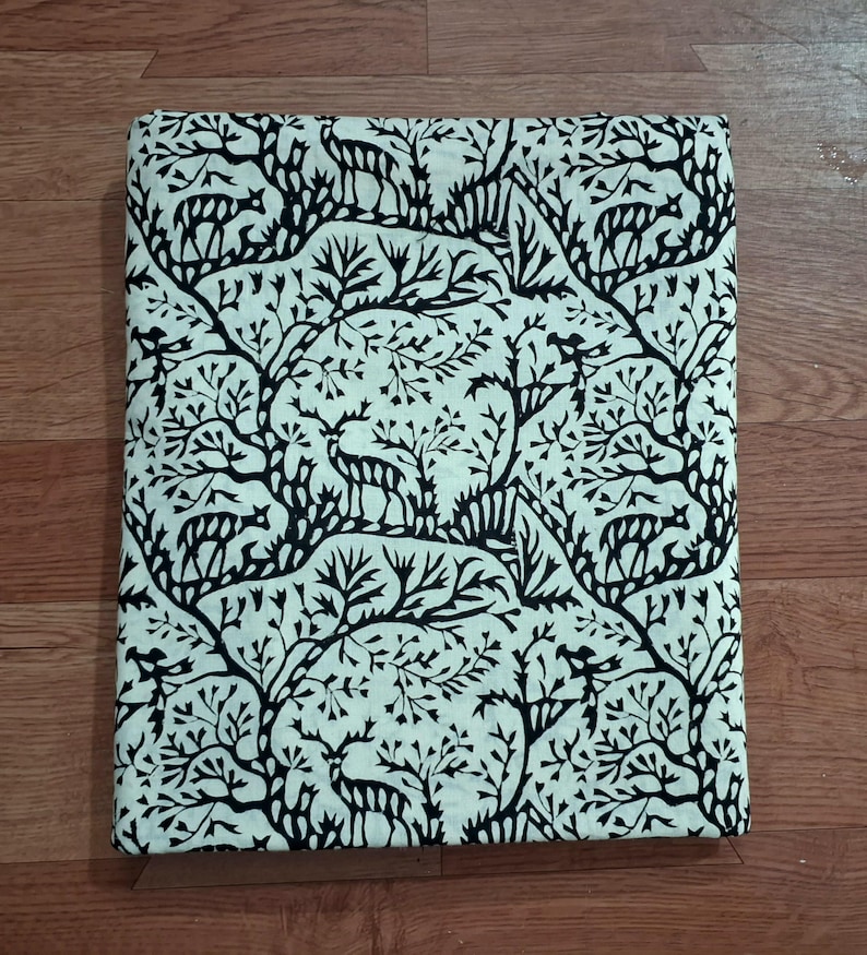 May include: A rectangular piece of fabric featuring a repeating pattern of black deer silhouettes interspersed among black stylized tree branches and foliage against a solid off-white background, folded into a neat, flat, rectangular shape.