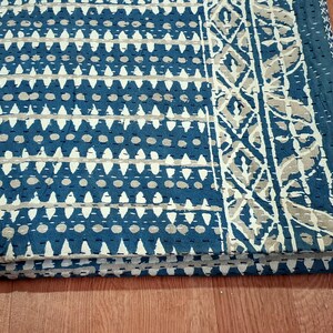 Puede incluir: Una colcha kantha azul y blanca con un patr&oacute;n geom&eacute;trico y un borde floral. La colcha est&aacute; doblada en tres partes y est&aacute; sobre una superficie de madera.