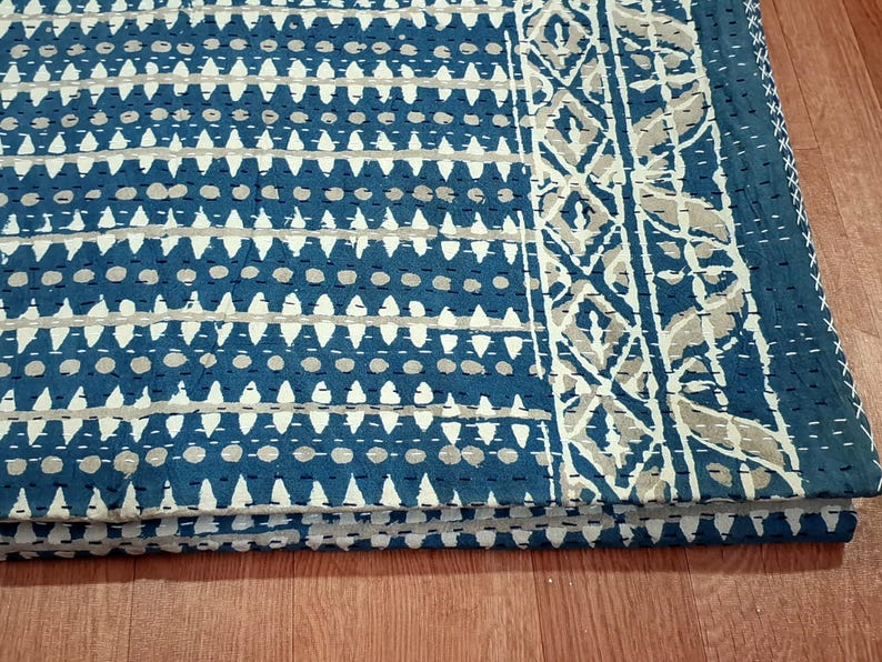 Puede incluir: Una colcha kantha azul y blanca con un patr&oacute;n geom&eacute;trico y un borde floral. La colcha est&aacute; doblada en tres partes y est&aacute; sobre una superficie de madera.