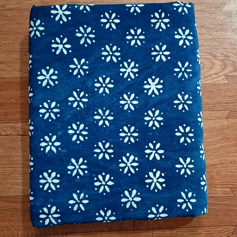 May include: A rectangular piece of fabric featuring a deep blue background with a repeating pattern of white, six-petalled floral shapes. The textile has a consistent, evenly spaced print across the entire visible surface, showcasing a simple, repetitive design.