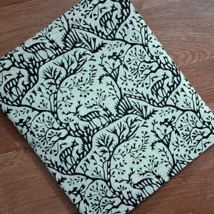 May include: A folded piece of fabric featuring a repeating black print on a light cream background. The pattern consists of stylized deer figures interspersed among intricate, branching tree designs that cover the entire surface area.