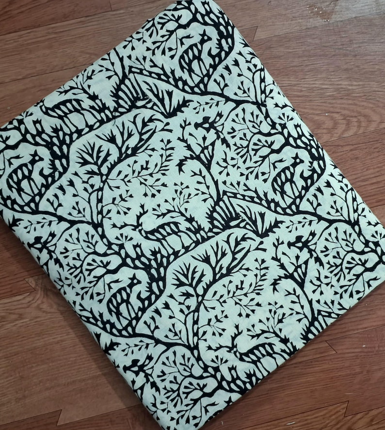 May include: A folded piece of fabric featuring a repeating black print on a light cream background. The pattern consists of stylized deer figures interspersed among intricate, branching tree designs that cover the entire surface area.