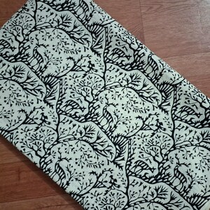 May include: A folded piece of fabric featuring a repeating pattern of black silhouettes of deer and trees against a cream-colored background. The design showcases stylized woodland creatures nestled among intricate, branching tree motifs.