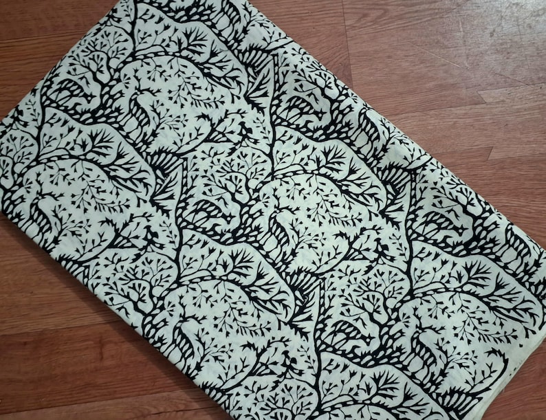 May include: A folded piece of fabric featuring a repeating pattern of black silhouettes of deer and trees against a cream-colored background. The design showcases stylized woodland creatures nestled among intricate, branching tree motifs.