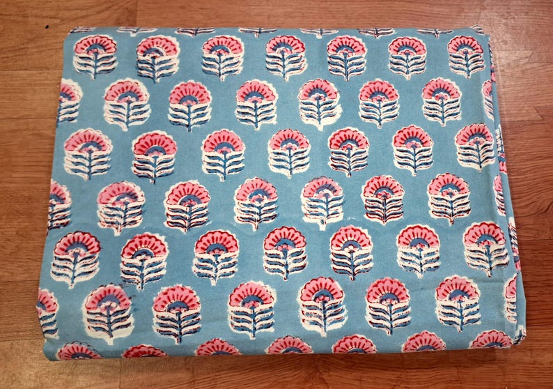 May include: A folded fabric with a repeating floral pattern. The flowers are pink and white with blue stems and leaves, set against a light blue background. The fabric is likely cotton or a similar material.